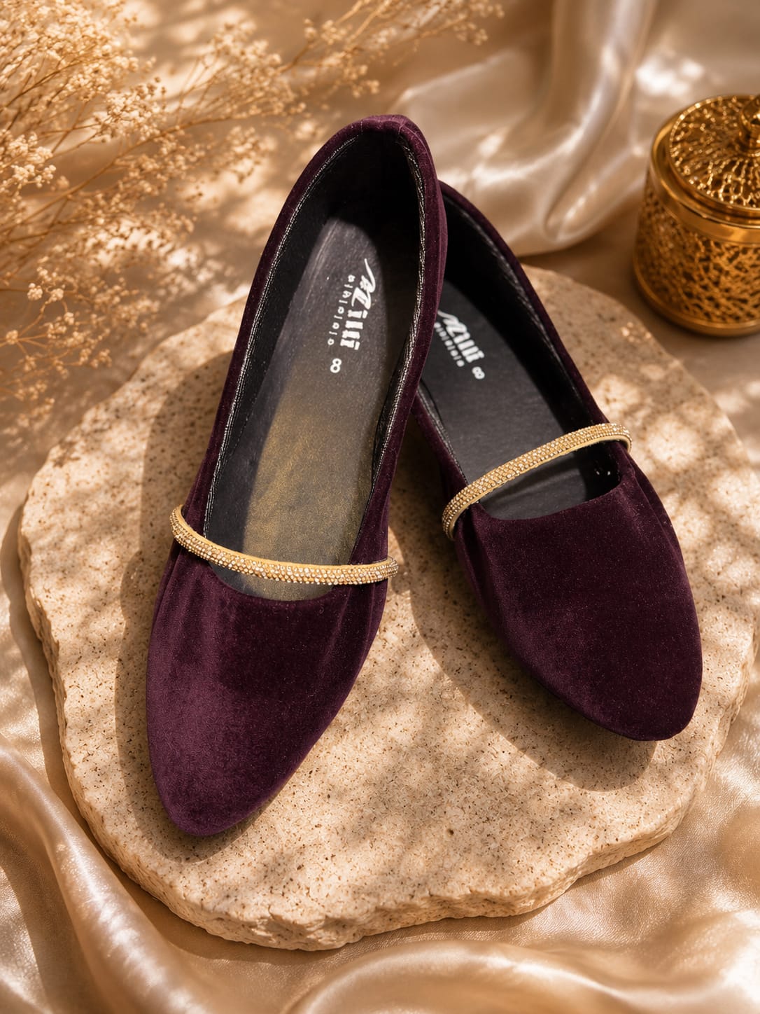 A pair of deep purple velvet slip-on flats with a thin gold chain detail across the top, displayed on a textured stone slab with gold satin fabric and a decorative gold container in the background.