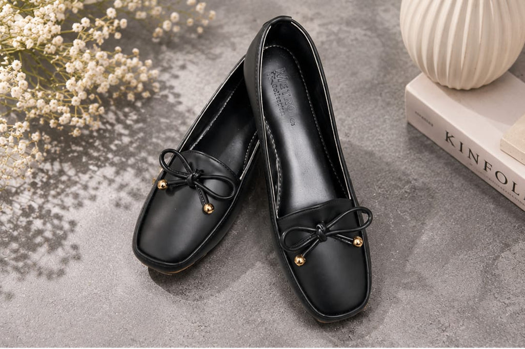 A pair of black leather loafers with a small black bow and gold bead detail on the toe, displayed on a textured gray surface with decorative flowers and a book.