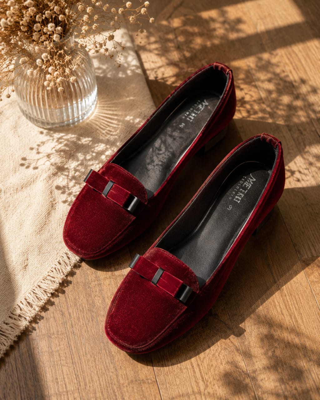 A pair of burgundy velvet loafers with a small black bow detail on the toe, displayed on a wooden floor next to a vase of dried flowers.