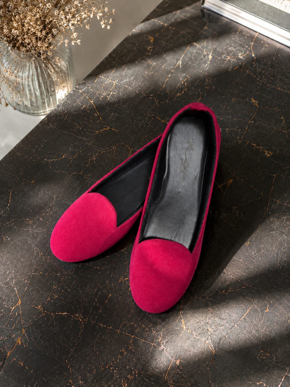 A pair of bright magenta velvet slip-on flats with black interiors, displayed on a dark marble surface with dramatic natural shadows.