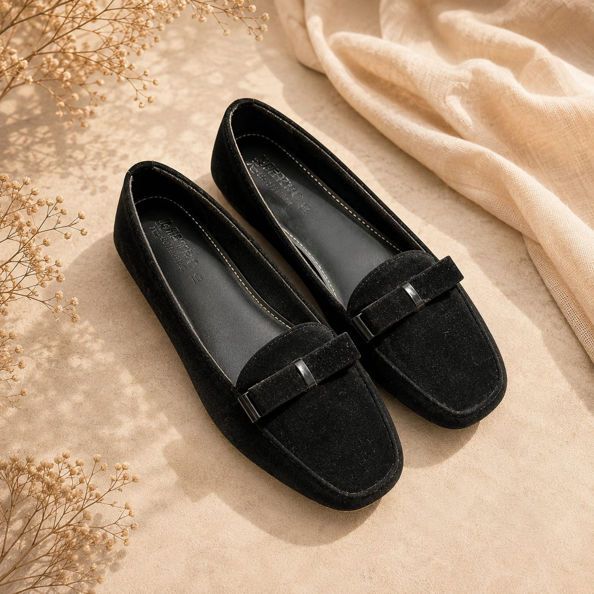 A pair of black suede loafers with square toes and a small metallic bow detail, styled on a beige textured surface with dried flowers and a cream fabric backdrop.