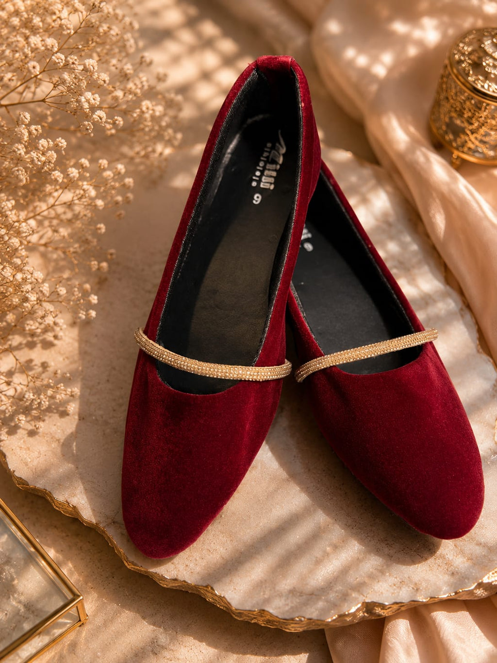A pair of deep burgundy velvet flat shoes with a thin gold rhinestone strap across the instep, displayed on a gold-rimmed stone slab with beige fabric and dried flowers.