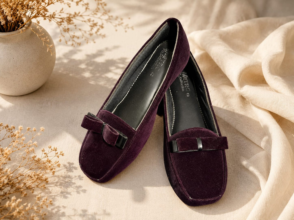 A pair of deep purple velvet loafers with bow details and black metal hardware, styled on a beige fabric surface with dried flowers and a cream-colored vase.
