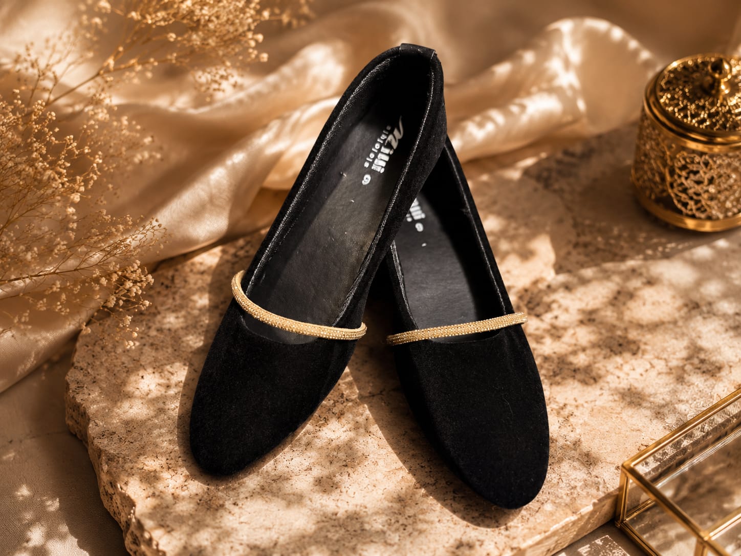 A pair of black suede flat shoes with a thin gold metallic trim across the vamp, displayed on a textured stone slab with draped champagne silk and dried floral accents.