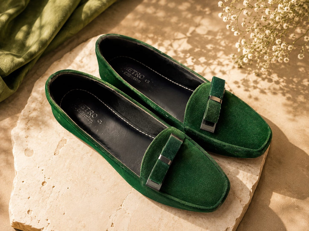 A pair of green suede loafers with a decorative strap and silver-tone hardware, displayed on a stone slab with a green fabric and dried flowers in the background.