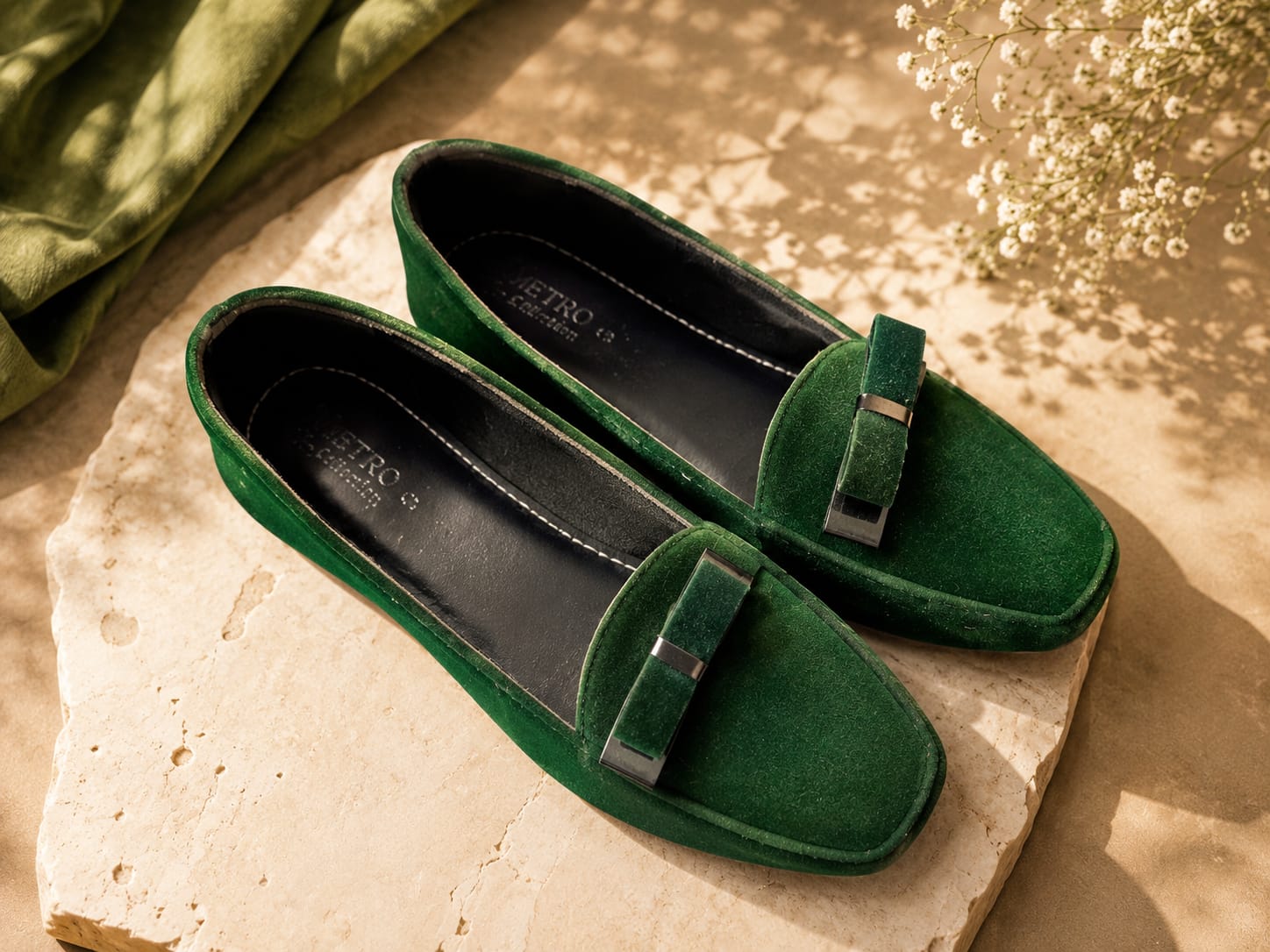 A pair of green suede loafers with a decorative strap and silver-tone hardware, displayed on a stone slab with a green fabric and dried flowers in the background.